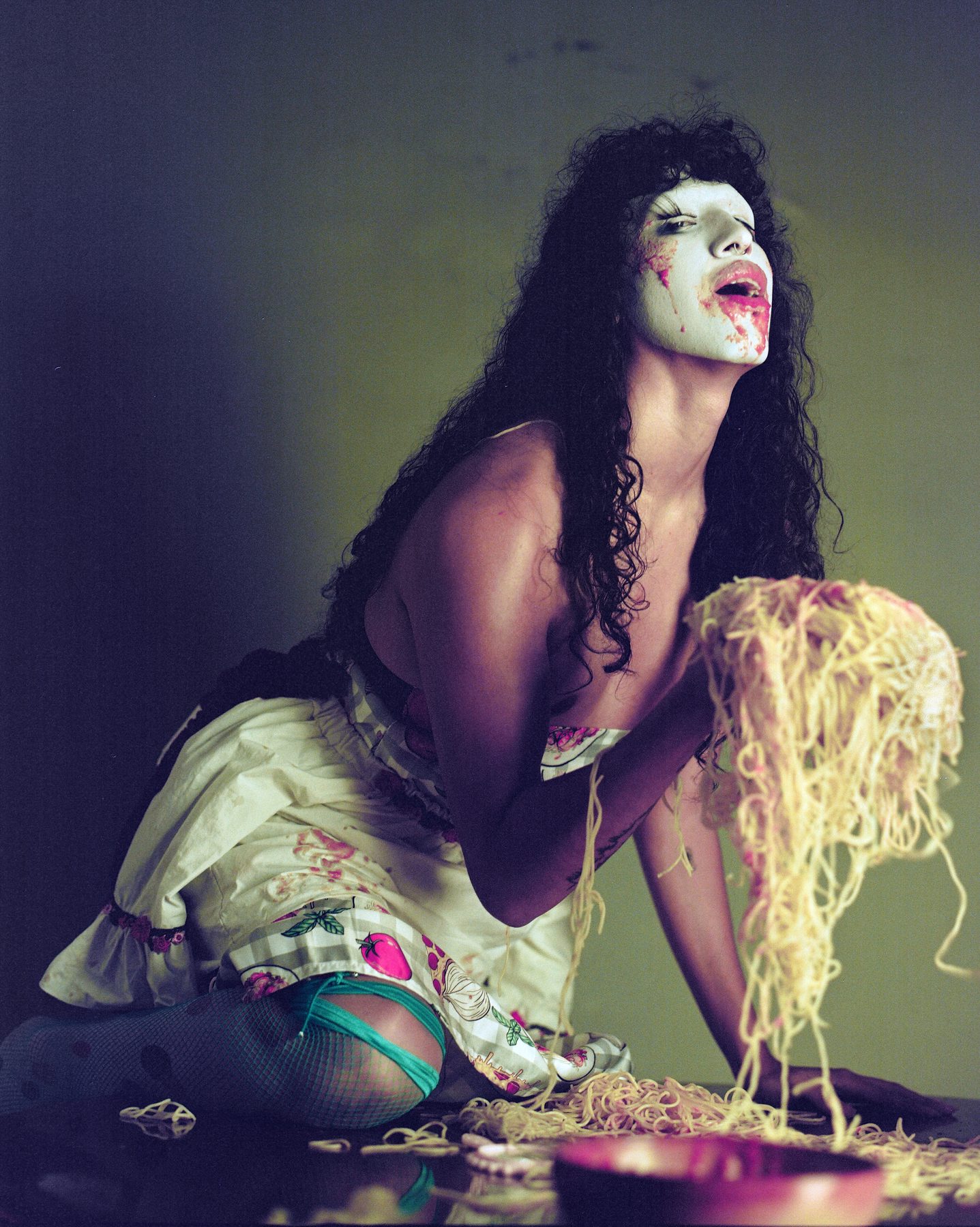 wiqi kneeling on a table in a white mask with smeared red lips and a gingham dress, lifting a mass of spaghetti