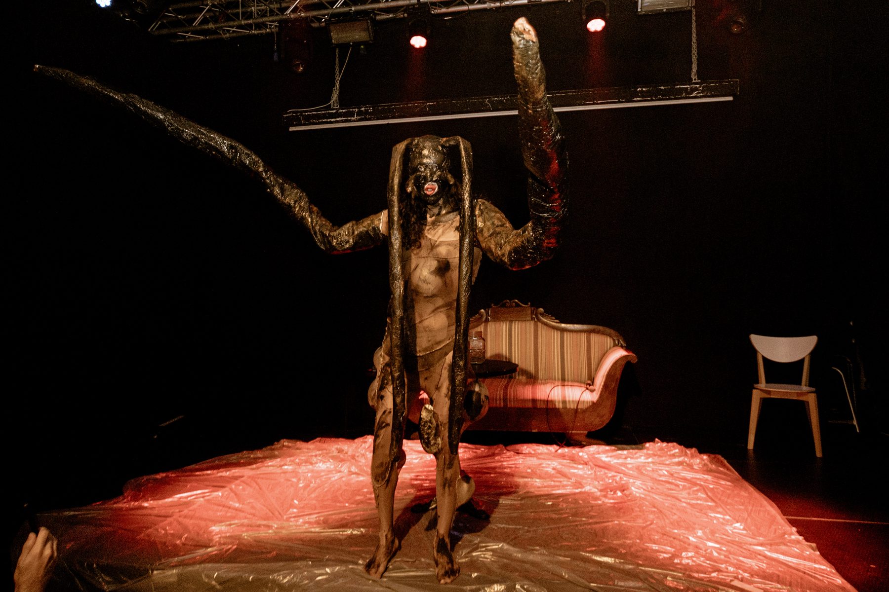 wiqi standing on red-lit plastic sheeting in a dark latex creature mask, with two long tentacle-like arms raised and a vintage sofa behind